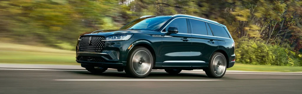 The image shows a black Lincoln Aviator SUV driving on a paved road surrounded by greenery.
