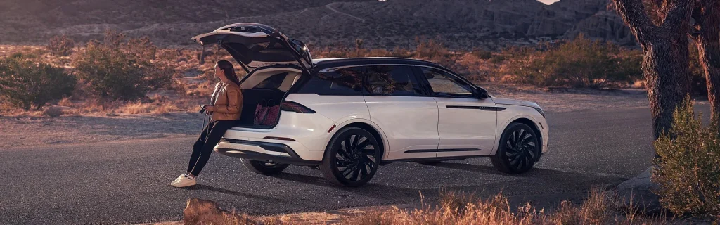 The image shows a woman sitting on the tailgate of a white Lincoln Nautilus SUV in a desert landscape.