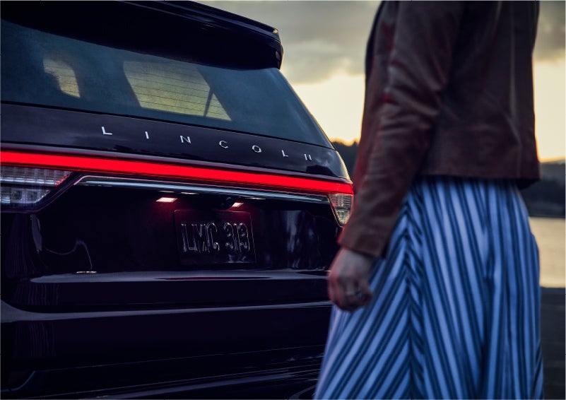 A person is shown near the rear of a 2023 Lincoln Aviator® SUV as the Lincoln Embrace illuminates the rear lights | Steet-Ponte Lincoln in Yorkville NY